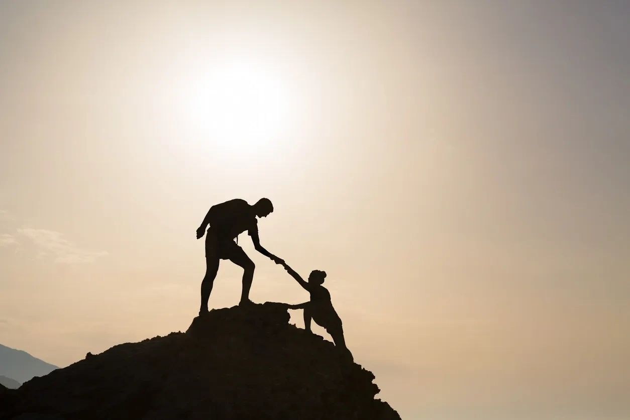 Silhouette of one figure helping another up a hilltop at sunset